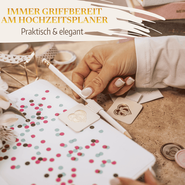 Eine Hand schreibt in ein Hochzeitsplaner-Notizbuch mit einem weißen Stift aus dem Mein kleines Hochzeitsbüro Stiftehalter - Schlaufe Standard. Buntes Konfetti, Anhänger, Klebeband und Blumen schmücken den beigen Schreibtisch. Text: Immer griffbereit am Hochzeitsplaner.