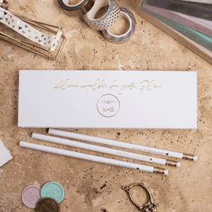 Eine Marmorplatte zeigt das Mein kleines Hochzeitsbüro Bleistift-Set - Lass uns 'ne Hochzeit planen mit goldenen Radiergummis, einer weißen Schachtel mit goldener Schrift, Klebebandrollen, Wachssiegeln und einer goldenen Heftklammer - ideal für die Planung Ihrer Hochzeit.
